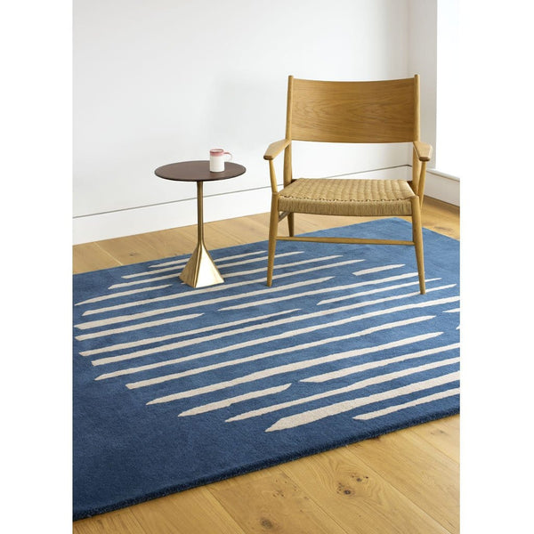 Island Blue Rug is an abstract lines patterned rug in two colours, blue and cream. The abstract lines pattern creates a circle shaped design within this rectangular wool rug. Pictured with chair and side table. 