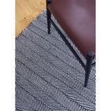Tibba Midnight Rug is a black and white monochrome rug, handmade from recycled plastic bottles. Herringbone style weave pattern. Pictured close up with chair.